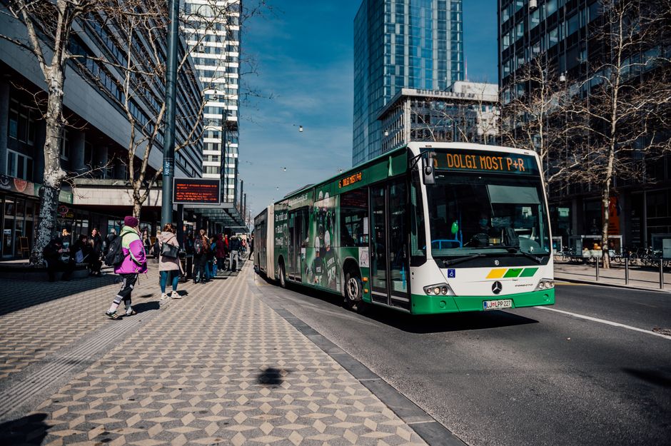 Mobilidade Urbana — Green bus in vibrant Ljubljana cityscape with pedestrians and modern buildings.