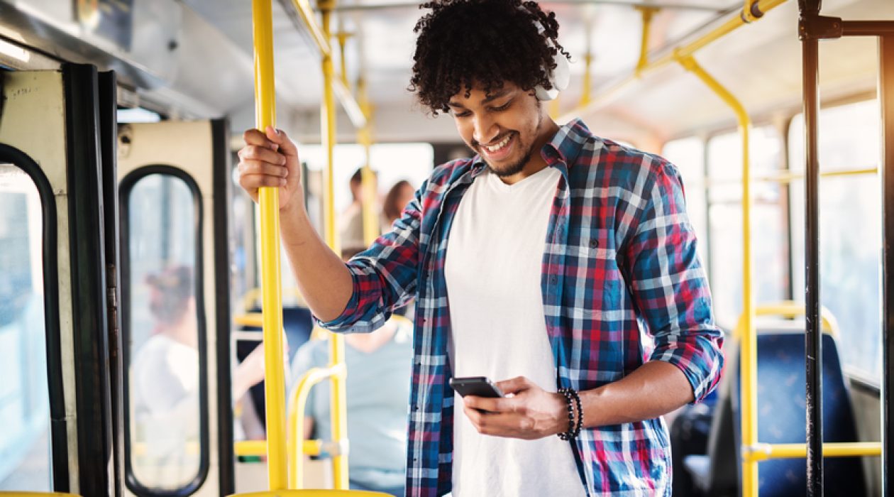 Young cheerful handsome man is enjoying the music during his ride and holding onto the bar while standing in a bus.