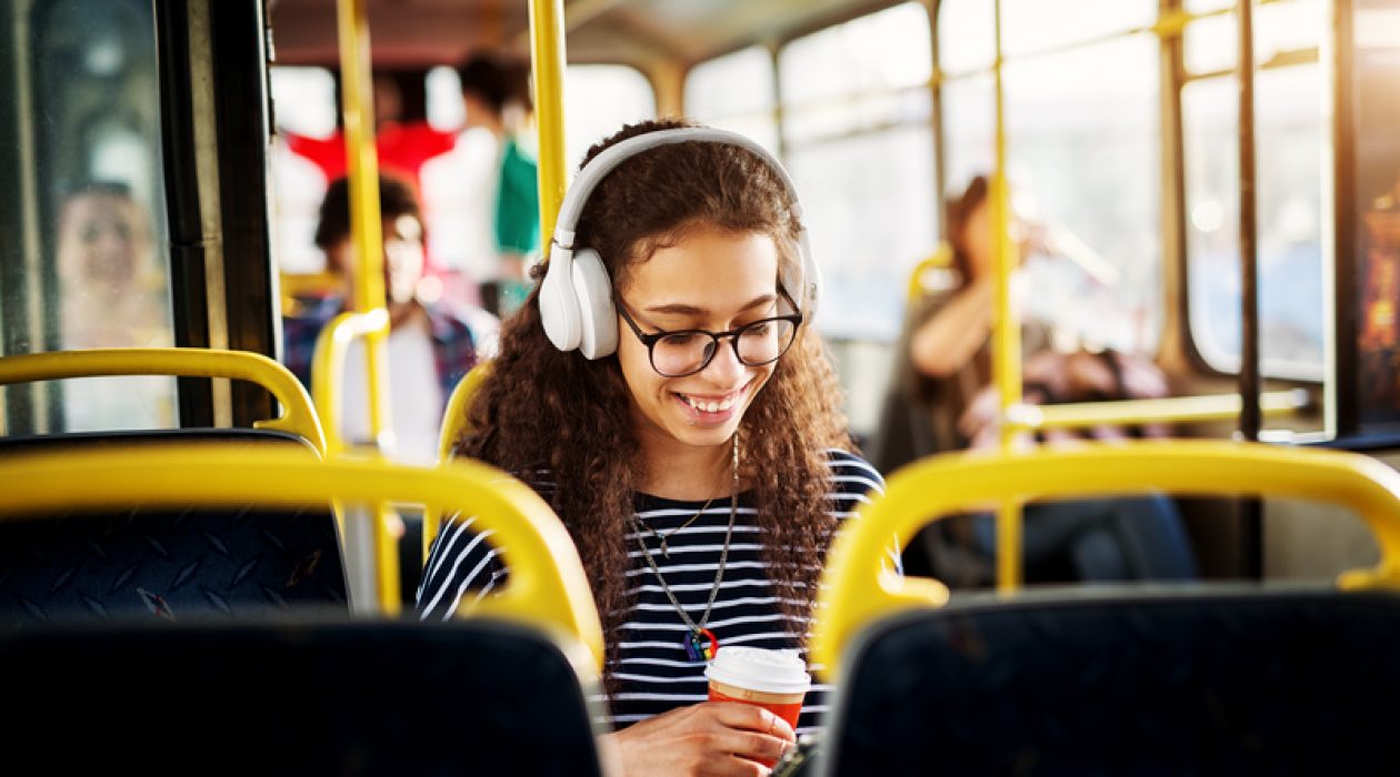 A gorgeous cheerful young woman with curly hair is sitting in the bus seat listening to music drinking coffee and using a tablet.